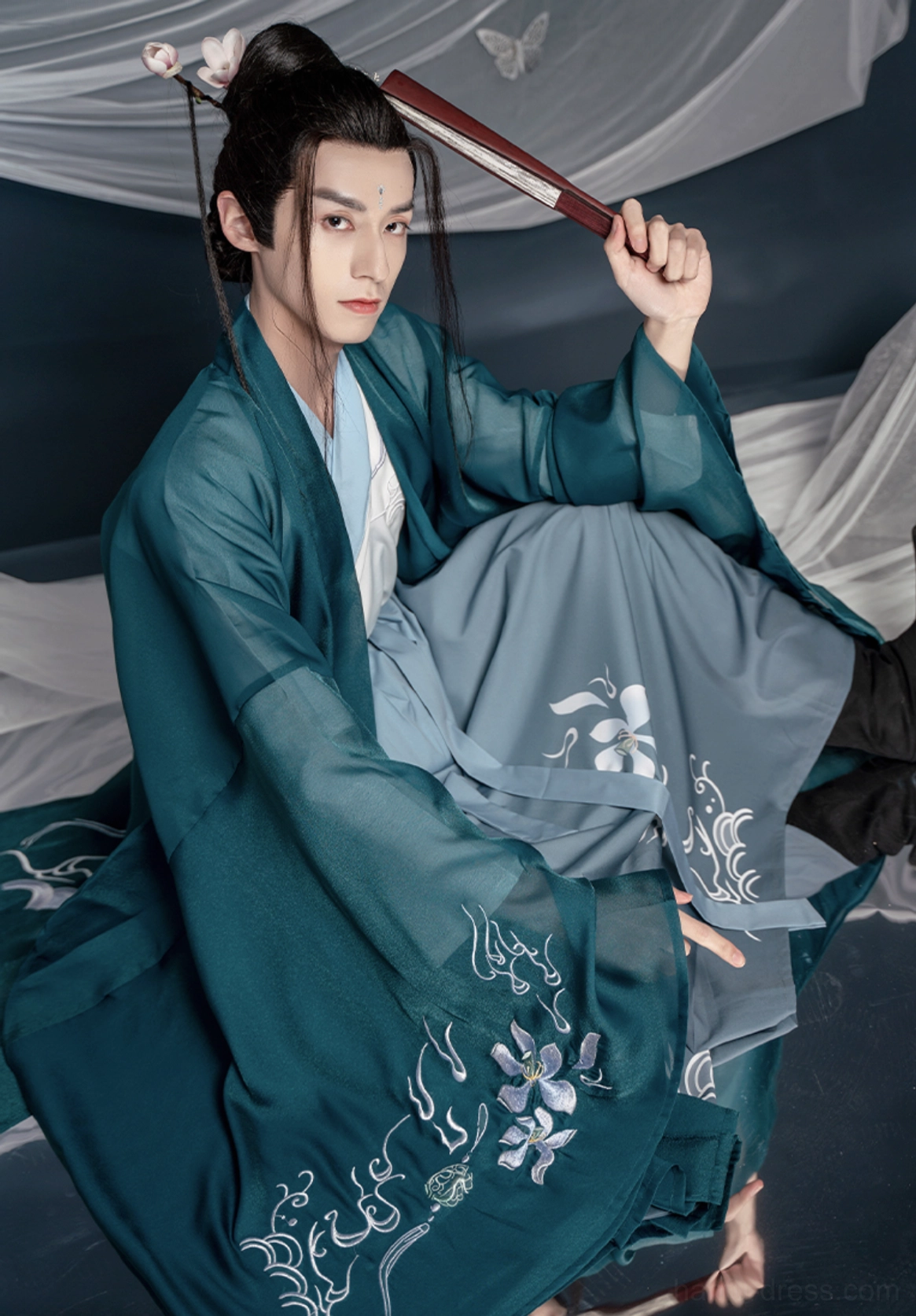 Side front sitting view of teal Wei Jin hanfu for men with embroidered outer coat, grey blue Qiyao skirt and traditional folding fan styling