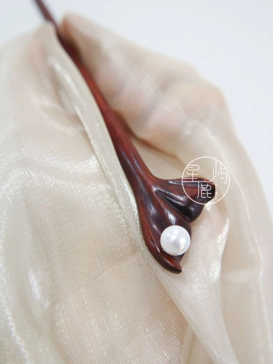 Top view of polished dark wooden Chinese hair stick with carved flower and white pearl-style ornament on silk fabric