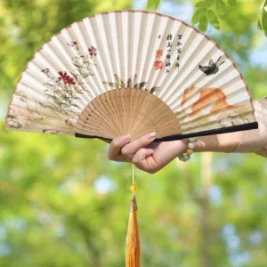 Cat Chinese folding hand fan with off-white printed surface, flowers, butterflies, Chinese calligraphy, smooth dark-edged bamboo ribs, and yellow tassel held outdoors against green foliage