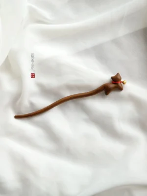 Full-length cat-shaped Chinese hair stick displayed on white linen, showing the long wooden shaft, lying cat ornament, red collar, and golden bell
