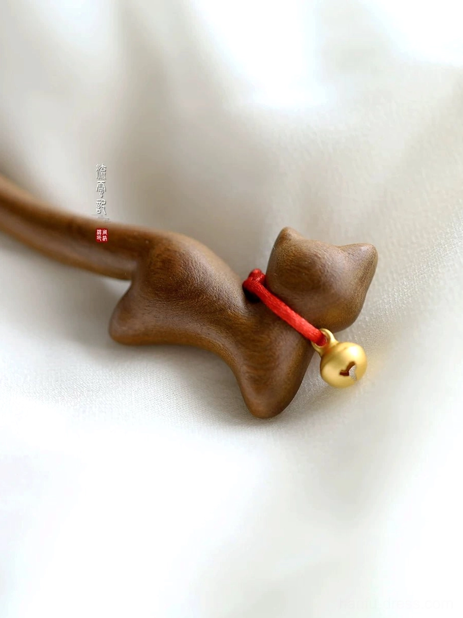 Top-down close-up of a cat-shaped wooden hair ornament showing the red collar and shiny golden bell against white linen fabric
