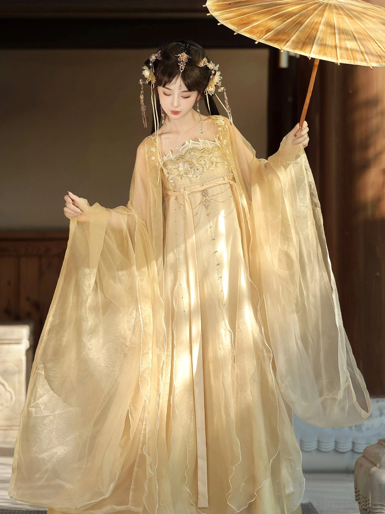 Champagne gold Tang Dynasty hanfu outfit shown from a right three-quarter angle with floral bust embroidery, dramatic ruffled organza sleeves, floor-length flowing skirt, and oil-paper umbrella beside a wooden building