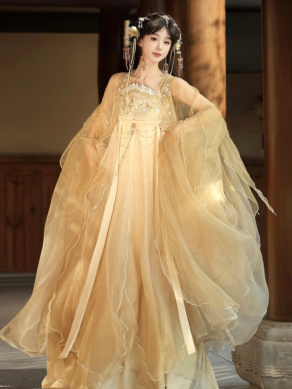 Champagne gold Tang Dynasty Qixiong Ruqun hanfu set shown from a front three-quarter view with sheer ruffled wide sleeves, floral embroidered bodice, flowing skirt, waist sash, and decorative waist chain in a traditional Chinese courtyard