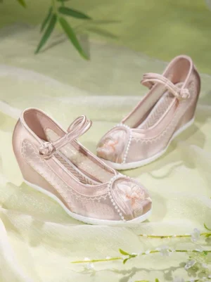 Champagne pink embroidered Chinese Hanfu shoes with floral toe embroidery, lace trim, frog-fastener ankle strap, and elevated wedge heel displayed on light yellow gauze with bamboo leaves