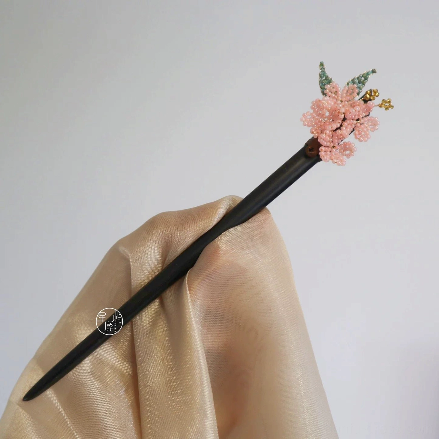Full product view of cherry blossom Chinese hair stick on champagne silk fabric with dark wooden stick, pink beaded blossom, green leaves, and gold-tone pendants
