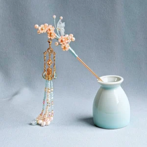 Cherry blossom tassel Chinese hair stick shown at an angle with peach floral clusters, faux pearls, gold beaded tassels, and light blue accents beside a porcelain vase