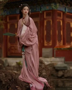 Full-body side view of cherry pink Warring States and Han Dynasty Hanfu straight-hem robe with wide sleeves, embroidered collar, decorative waist pendant, and green silk fan in an ancient Chinese courtyard
