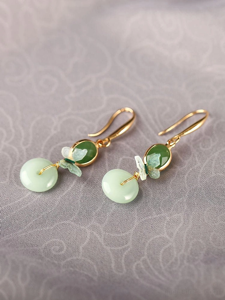 Pair of Chinese butterfly jade drop earrings arranged on embroidered fabric showing gold hardware, green oval stones, enamel butterflies, and round peace buckle pendants