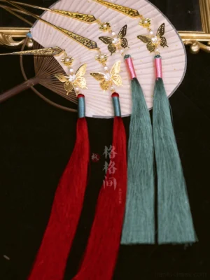 Red and green Chinese tassel hair pins with gold-tone butterfly ornaments displayed beside a Chinese folding fan