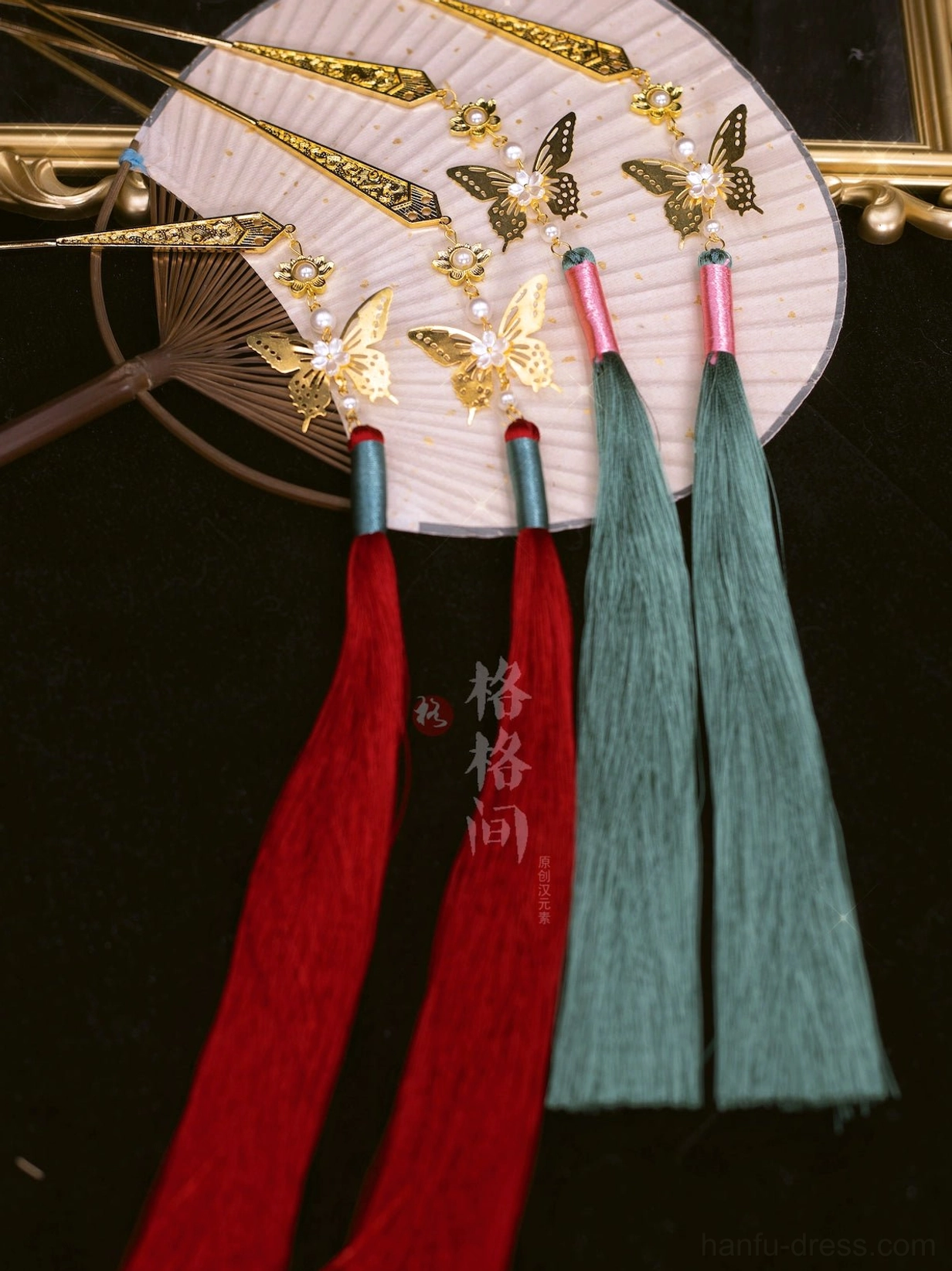 Red and green Chinese tassel hair pins with gold-tone butterfly ornaments displayed beside a Chinese folding fan