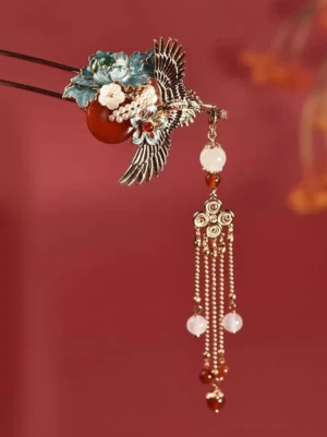 Chinese crane tassel hair stick close-up with gold-tone metal, blue floral enamel-style details, red center accent, bead pendants, and flowing chain tassels on dark red background