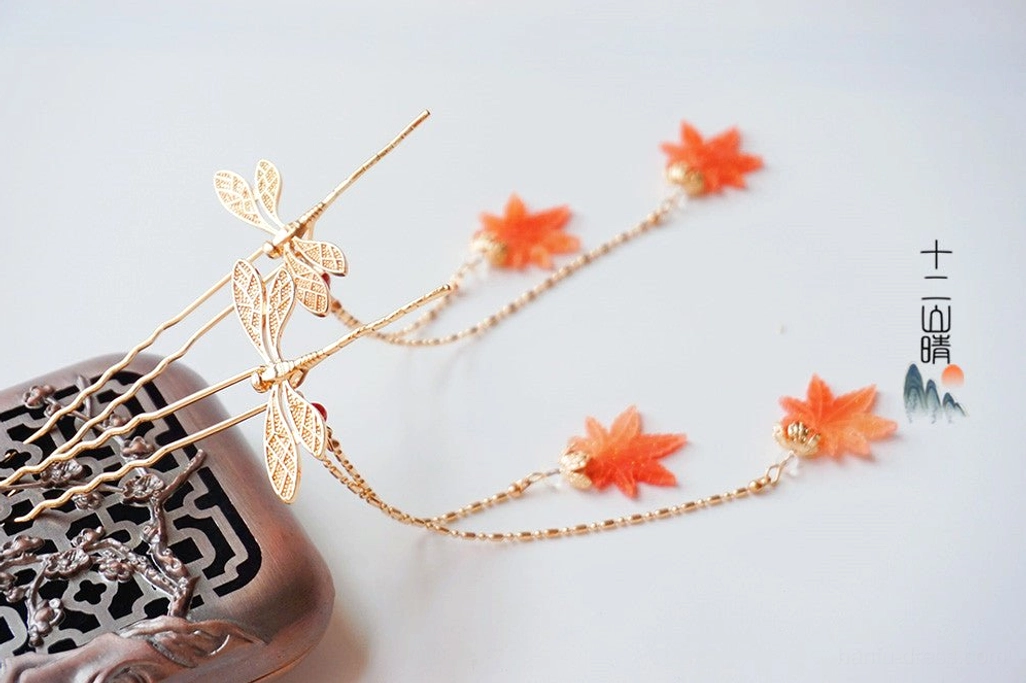 Pair of Chinese dragonfly maple leaf hair pins with orange leaf charms and delicate gold chain tassels displayed on a carved wooden coaster