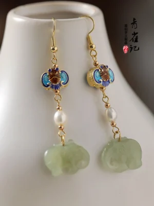 Chinese elephant dangle earrings with light green jade-tone elephant charms, cloisonne bat ornaments, freshwater pearls, and gold-tone hooks displayed beside a white porcelain vase