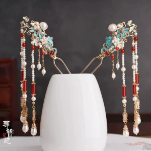 Pair of traditional Chinese enamel tassel hairpins with blue floral ornaments, pearl accents, red agate details, gold-toned metal, and long hanging pearl pendants displayed on a white ceramic vase