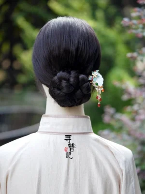 Woman wearing a Chinese fan-inspired tassel Hanfu hairpin in a braided low bun, showing the elegant beaded pendants and decorative ornament against a garden background