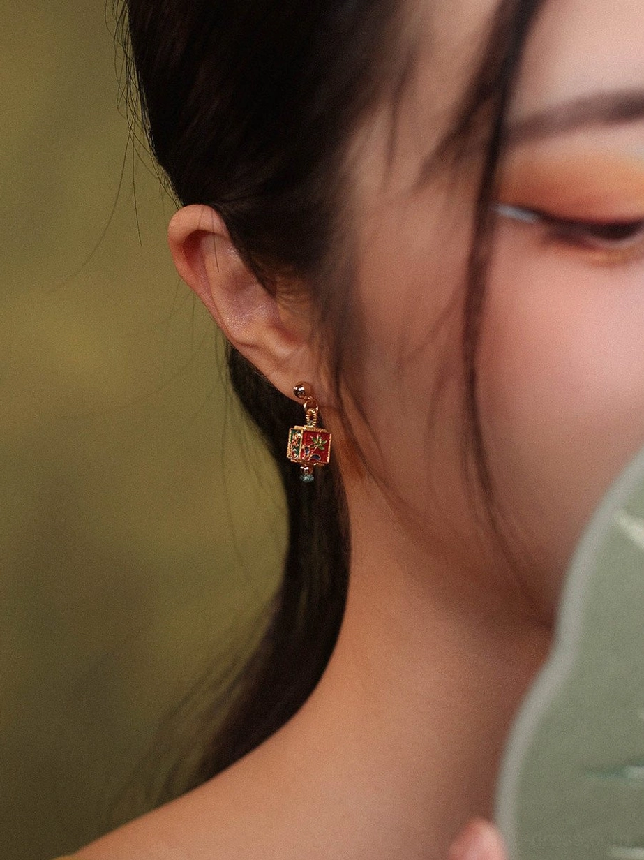 Close-up wearing view of Chinese floral cube drop earrings with gold-tone setting and blue bead accents paired with a green folding fan