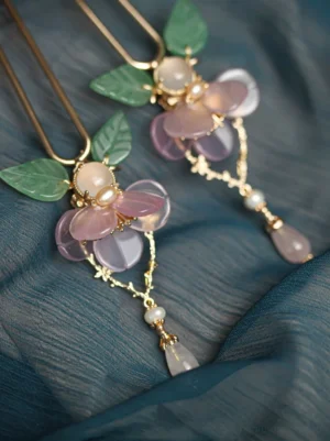Chinese floral hair pin displayed on dark blue fabric with pink-purple petals, green leaves, gold double prongs, pearl beads, glass-like drops, and decorative tassel