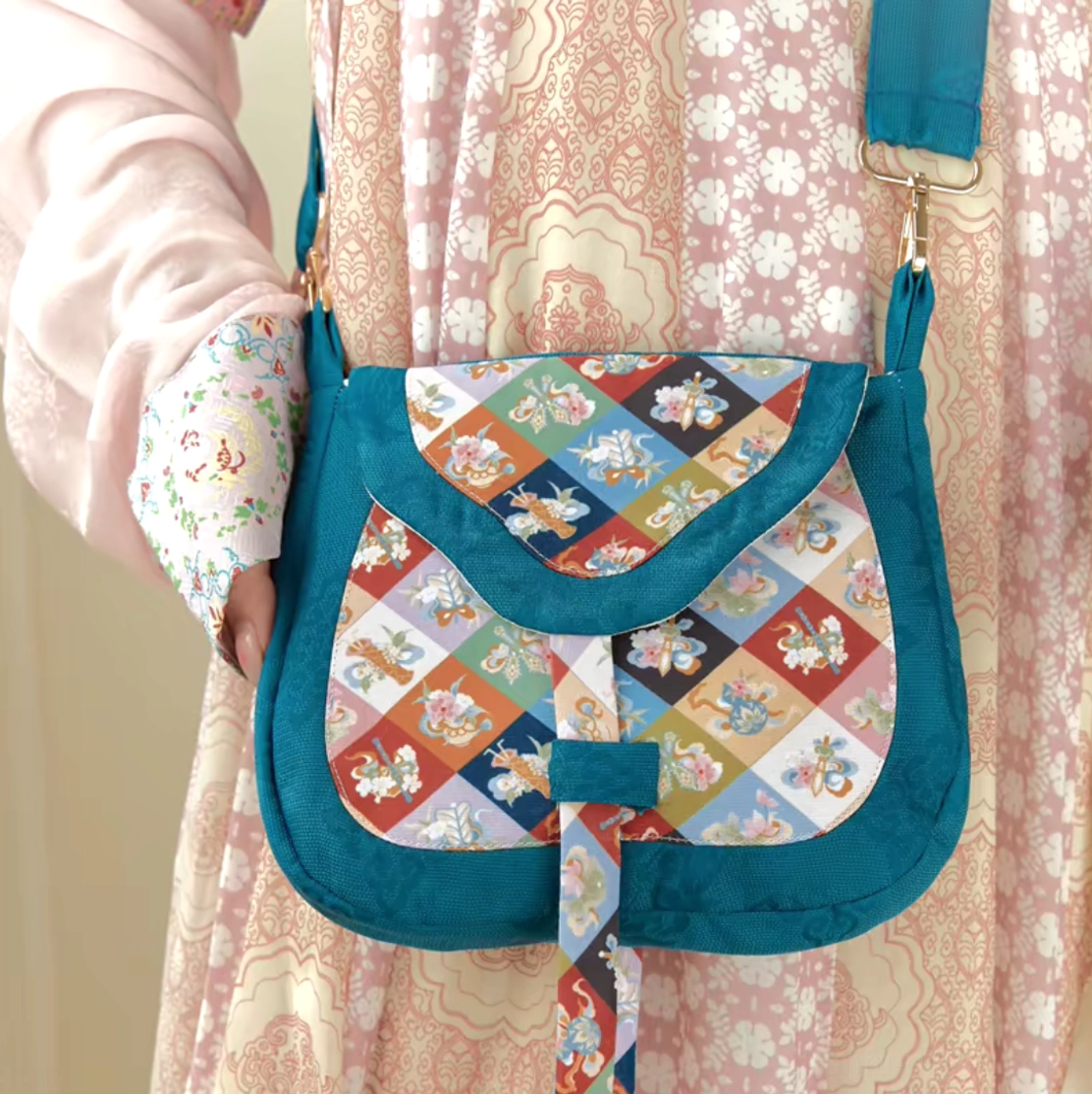 Chinese floral patchwork crossbody bag styled with a pink floral Hanfu skirt, featuring teal border trim, diamond motifs, adjustable strap, and gold buckles