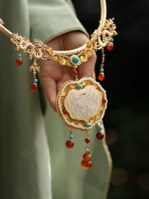Close-up of a traditional Chinese gold longevity lock necklace held by a hand in a light green Hanfu sleeve, showing gold dragon filigree, turquoise and red agate accents, a carved white jade pendant and pearl trim