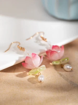 Chinese lotus dangle earrings with gradient pink flower charms, gold ear hooks, green accents, and freshwater pearls on a beige tabletop