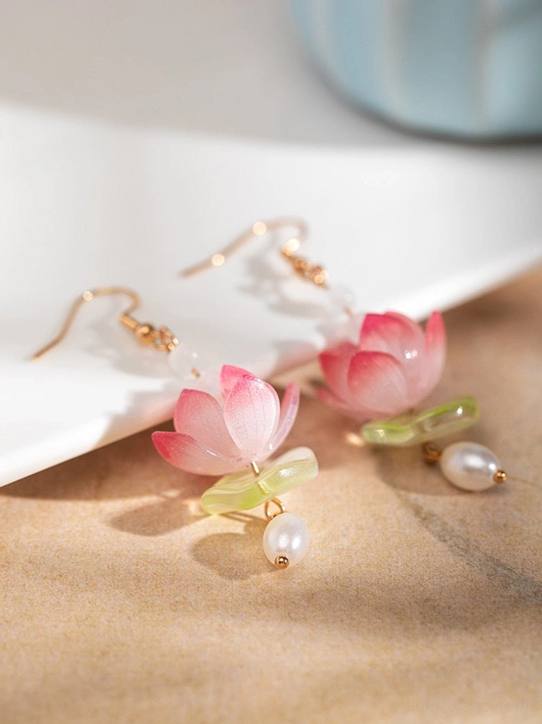 Chinese lotus dangle earrings with gradient pink flower charms, gold ear hooks, green accents, and freshwater pearls on a beige tabletop