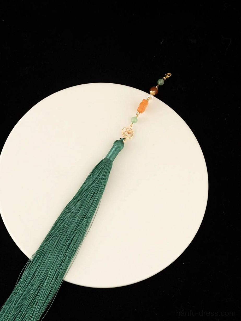 Close-up side view of the green silk tassel pendant on a Chinese-style necklace, with a warm orange bead and gold-tone fittings displayed on a white plate