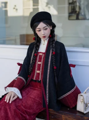 Seated female model wearing a modern Chinese New Year Hanfu outfit with a black cropped jacket, gold braided trim, red inner top, festive braided hair and black beret