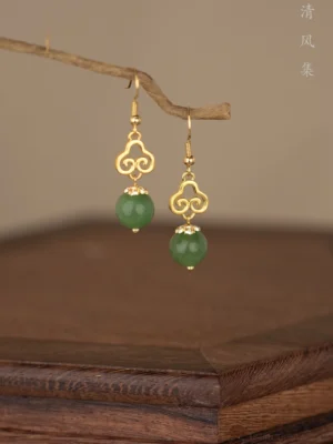 Close-up of traditional Chinese Ruyi jade drop earrings with 925 silver hooks, gold openwork auspicious motifs, dark green round pendants, and delicate rhinestone accents displayed on a wooden branch