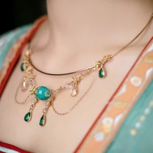 Woman wearing a gold-tone Chinese style yingluo collar necklace with teal center bead, green teardrop gems, clear pendants, floral details, and layered tassel chains with Hanfu