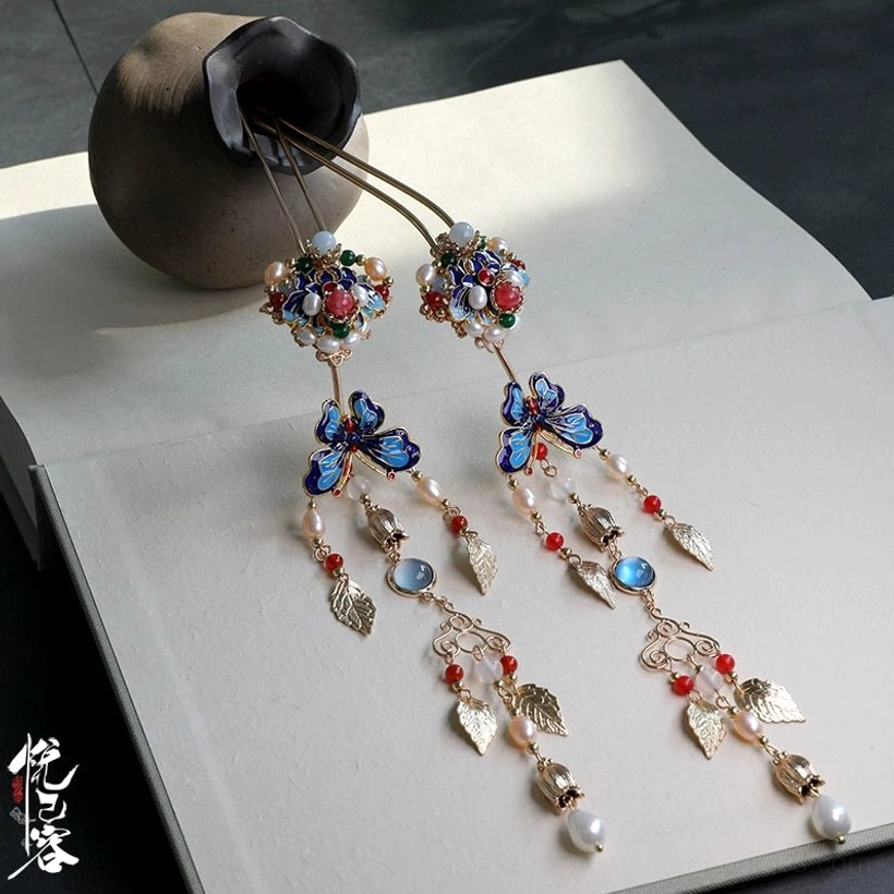 Chinese tassel hair pin with cloisonne butterfly, red beads, golden leaf charms, enamel flower, and pearl pendant displayed beside a ceramic vase