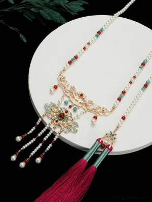 Side flat lay view of a Chinese tassel hanfu necklace with pearl-style chain, red and turquoise beads, gold cloisonne pendant, and rose-red tassel on a white display plate
