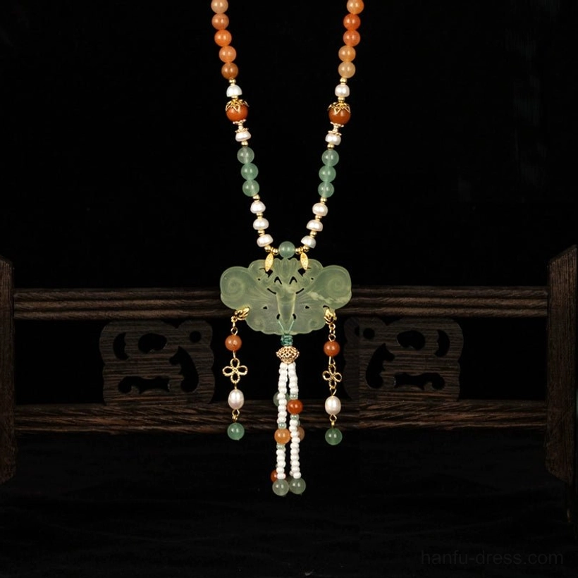 Three-quarter front view of a Chinese tassel necklace on a vintage wooden jewelry stand, featuring gold-tone floral embellishments, a green pendant, warm beads, and detailed hanging decorations