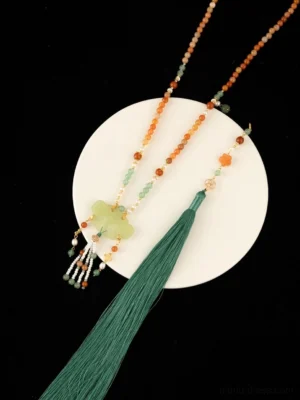 Flat lay close-up of a Chinese tassel necklace displayed on a white ceramic plate, showing orange beads, a green decorative charm, pearl accents, and a long green tassel