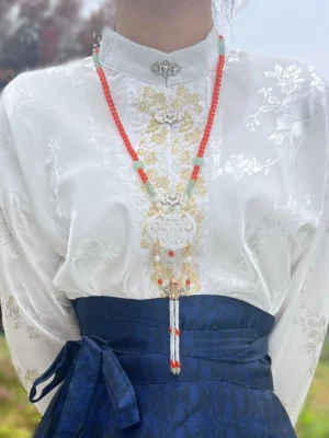 Woman wearing a Chinese style yingluo necklace with red beads, silver floral ornaments, jade details, and front tassel over a white Hanfu top and dark blue mamian skirt