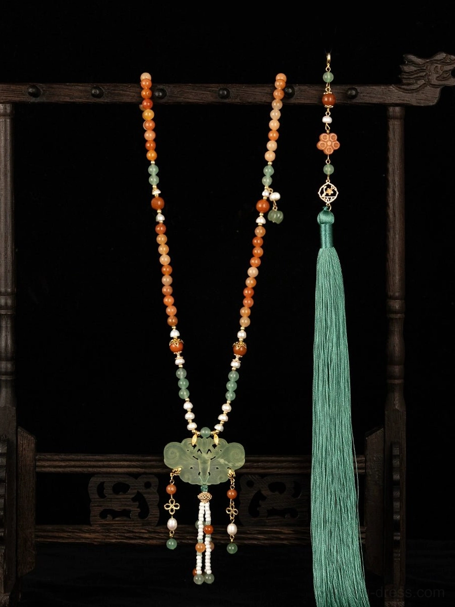 Full front view of a traditional Chinese yingluo tassel necklace on a vintage wooden jewelry stand, featuring warm-toned beads, a green ornamental pendant, and a long flowing silk tassel against a black background