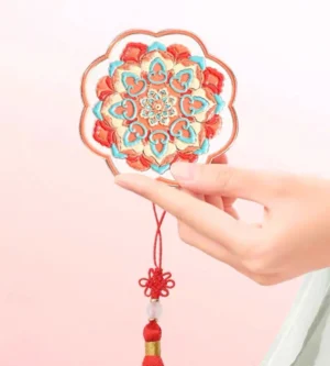 Chinese hand mirror held in a hand against a pink background with Hanfu sleeve, showing the raised cloisonne-style lotus floral pattern and traditional red tassel