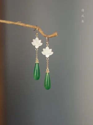 Classic jade lotus drop Chinese earrings displayed as a symmetrical pair on a dry twig, featuring carved white shell lotus charms, gold-tone chains, and deep green teardrop pendants