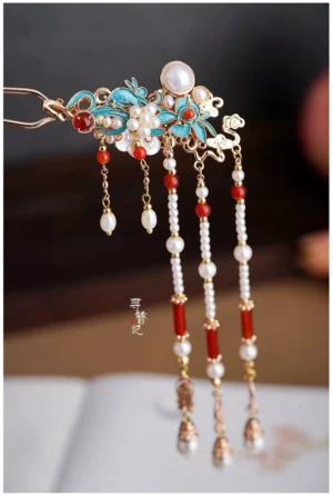 Close-up of a classical Chinese tassel hairpin with blue enamel flowers, a large pearl, red agate accent, small pearls, and layered hanging tassels beside an open book