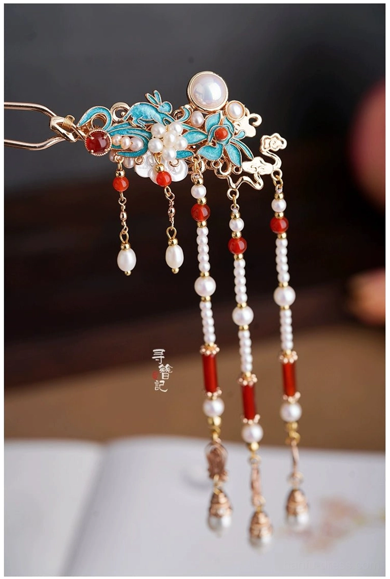 Close-up of a classical Chinese tassel hairpin with blue enamel flowers, a large pearl, red agate accent, small pearls, and layered hanging tassels beside an open book