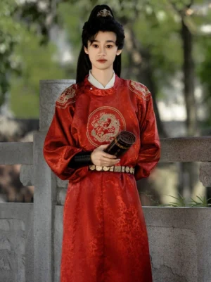 Front view of male model wearing a red Tang style round-collar hanfu robe with gold dragon motif and decorative belt on a traditional stone bridge