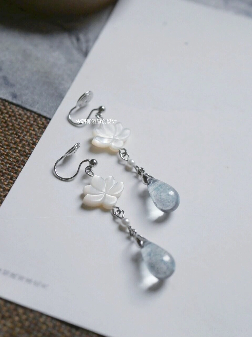 Two pairs of Chinese lotus dangle earrings with white carved flower pendants blue glass drops and silver hooks on woven fabric background