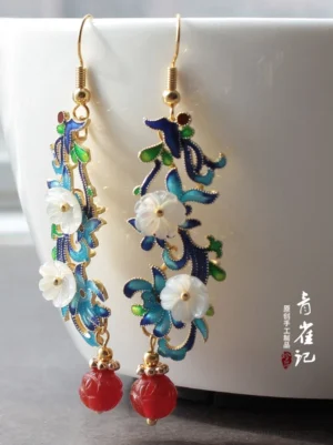 Antique-inspired cloisonne flower drop Chinese earrings with gold hooks, blue-green floral enamel details, white carved shell flowers, and red bead pendants displayed beside a white ceramic mug