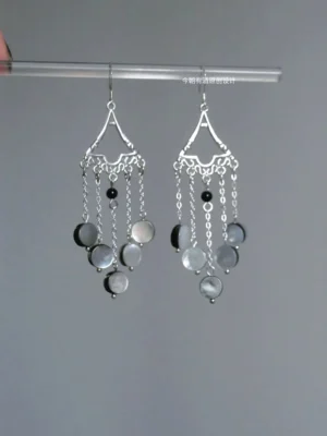 Close-up Chinese Hanfu tassel earrings hanging from a clear display rod, showing hollow carved silver ear tops, gray shell pendants, black accent beads, and flowing chain tassels