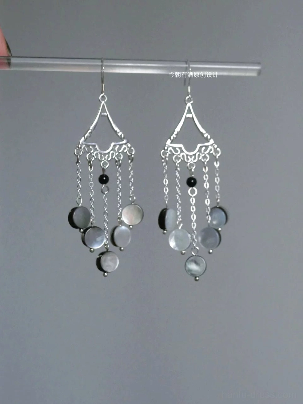Close-up Chinese Hanfu tassel earrings hanging from a clear display rod, showing hollow carved silver ear tops, gray shell pendants, black accent beads, and flowing chain tassels