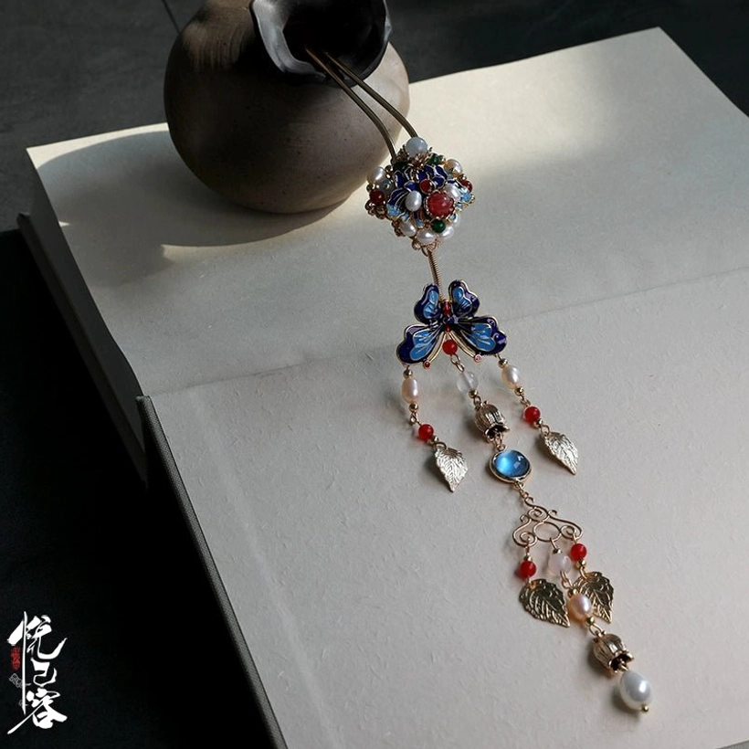 Close-up Chinese ornamental hair pin showing enamel flower, cloisonne butterfly, red agate-style beads, golden leaf charms, tassel, and pearl pendant