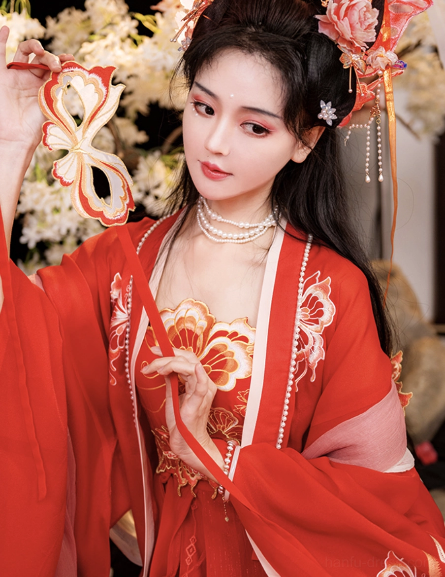 Close-up of a Tang-style red bridal hanfu bodice with golden lotus and butterfly embroidery, pearl neckline trim, pink sleeve accents, layered pearl necklace, and traditional floral accessories