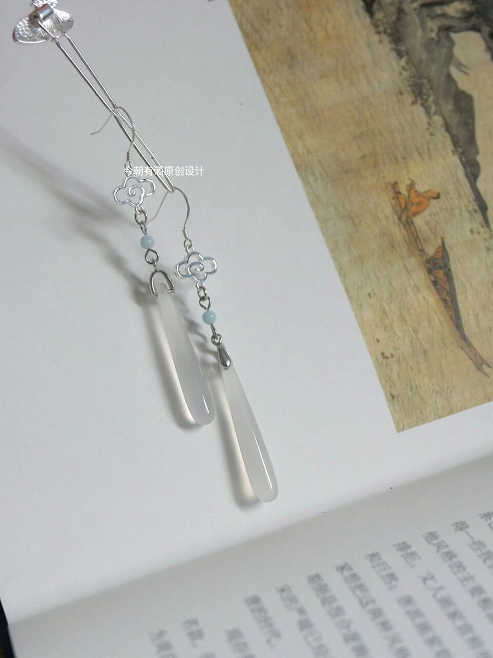 Cloud motif Hanfu drop earrings with white agate pendant and detailed ear hook structure placed on an ancient Chinese book with traditional ink painting