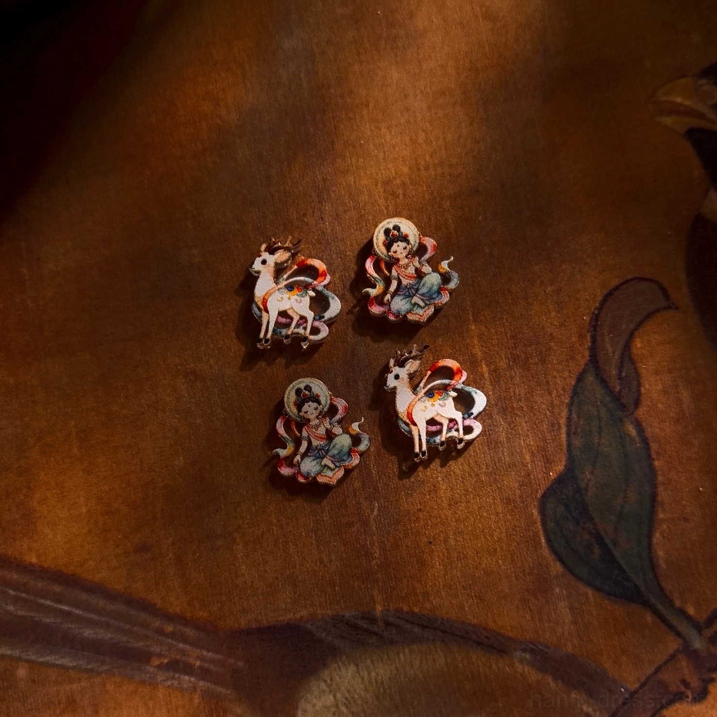 Two pairs of colorful Dunhuang inspired stud earrings with Apsara and Nine-Colored Deer motifs arranged on a retro Chinese wooden backdrop
