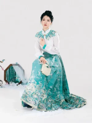 Model seated in a complete Chinese Hanfu outfit with jacquard long sleeve top cloud collar and flowing teal Mamian skirt beside oriental decor