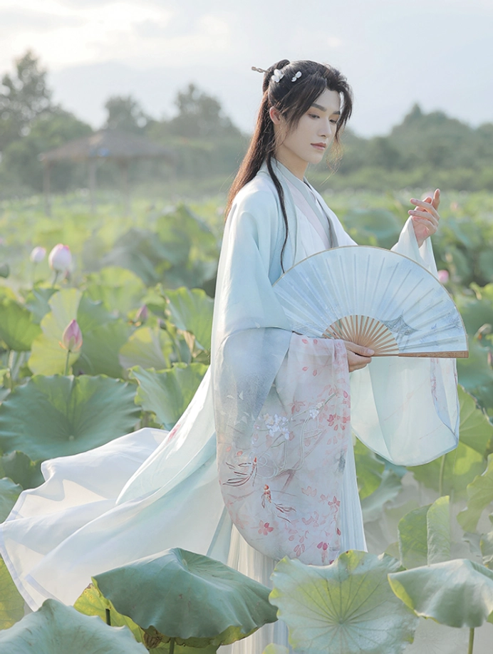 Three-quarter front view of the complete Cloud Pavilion hanfu set for men with soft cyan and ivory layers, embroidered sleeve details, flowing robe, and folding fan in a lotus pond setting